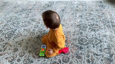 baby boy in orange suit sitting on the floor playing with toy phone. kid pressing the buttons on the toy. top view.