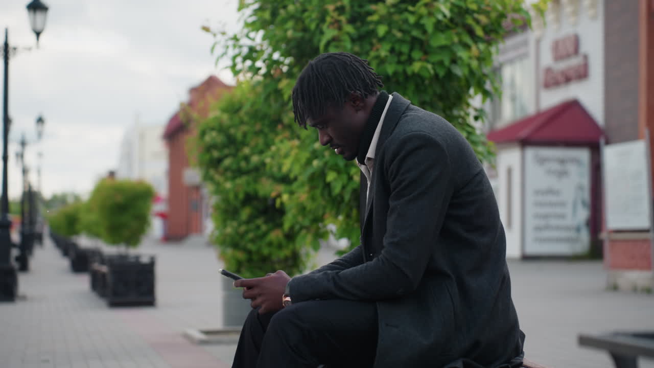 Male adult walking on boulevard using phone while holding bag, dressed in stylish coat, showing confident posture and focus, surrounded by trees and streetlights in peaceful urban setting