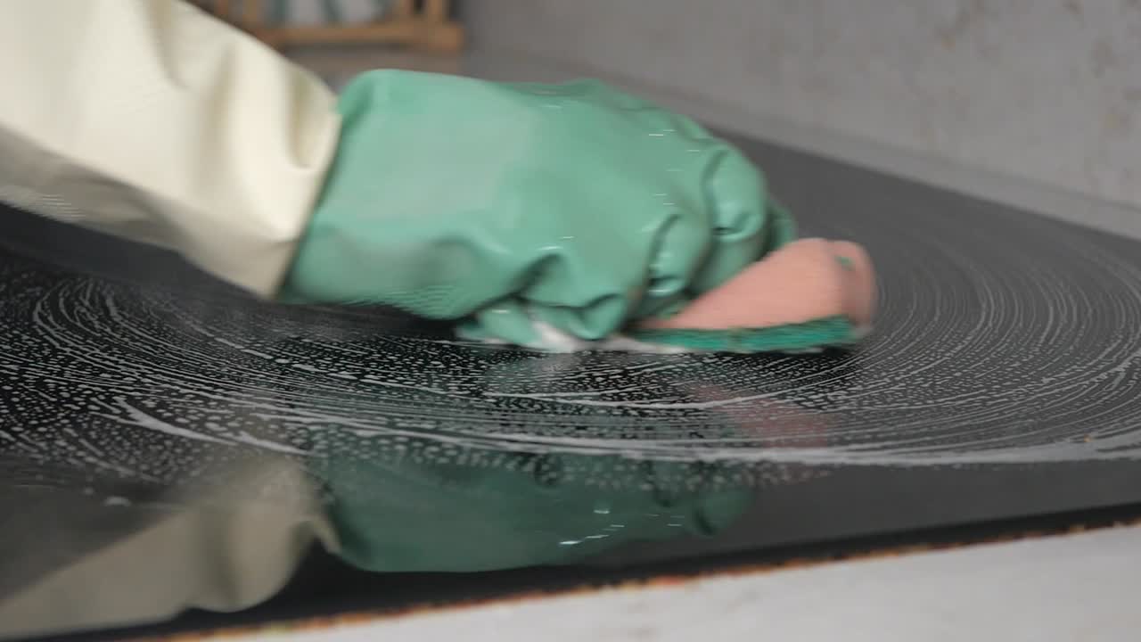 Hand in grünem Handschuh reinigt Induktionskochfeld mit Schwamm.