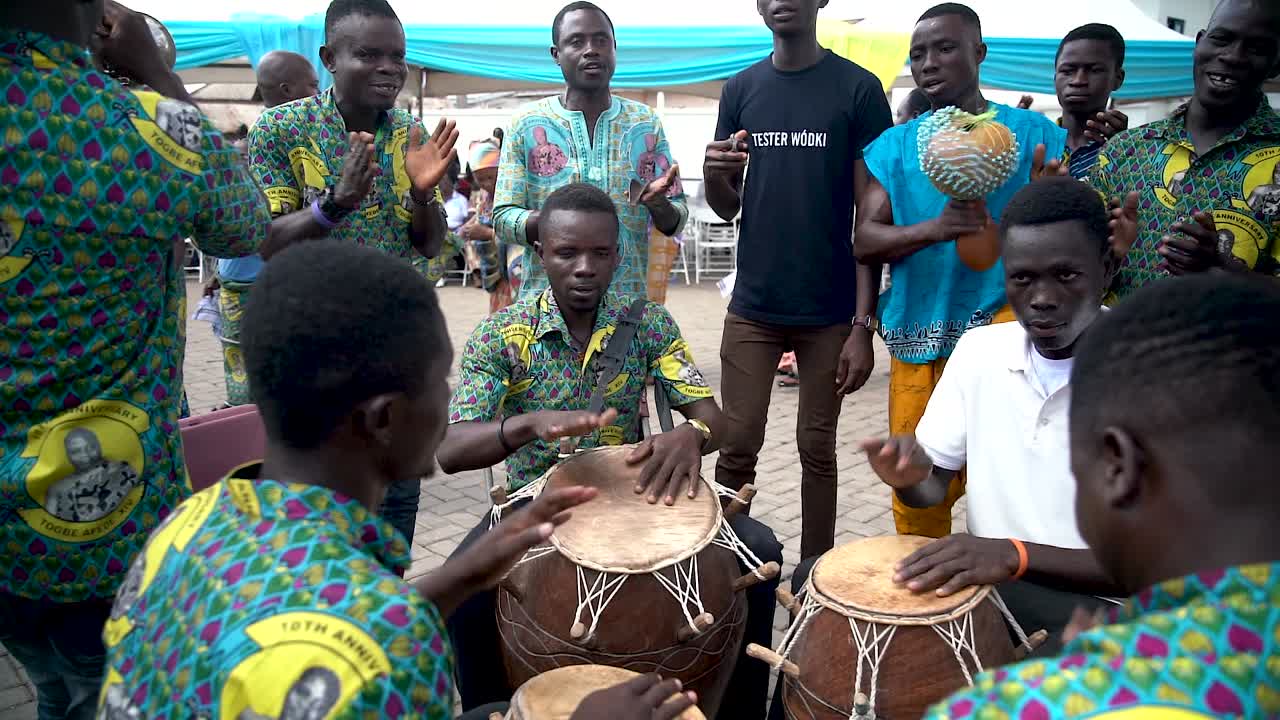 A music group of drummers perform while the camera tilts up to reveal other musicians dancing and playing.