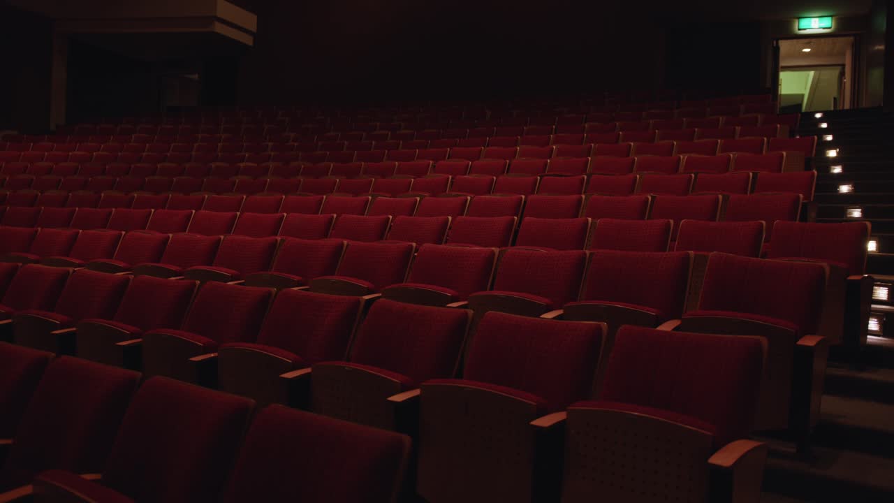 Wide view of tiered rows of empty red theater seats inside a dimly lit auditorium. Slow motion view