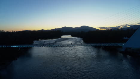 Empuje-Aéreo-Lento-Hacia-El-Puente-Del-Reloj-De-Sol-Mientras-La-Gente-Camina-A-Través-De-él-Al-Atardecer-Sobre-El-Río-Sacramento-En-Redding,-California