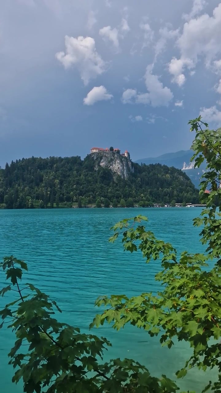 Vista verticale del Lago di Bled e del Castello di Bled in una soleggiata giornata estiva, con acqua turchese e punto di riferimento.