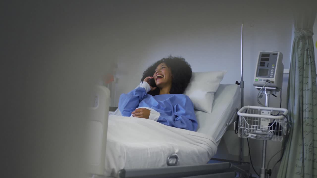 paciente afroamericana acostada en una cama de hospital hablando en un teléfono inteligente y sonriendo