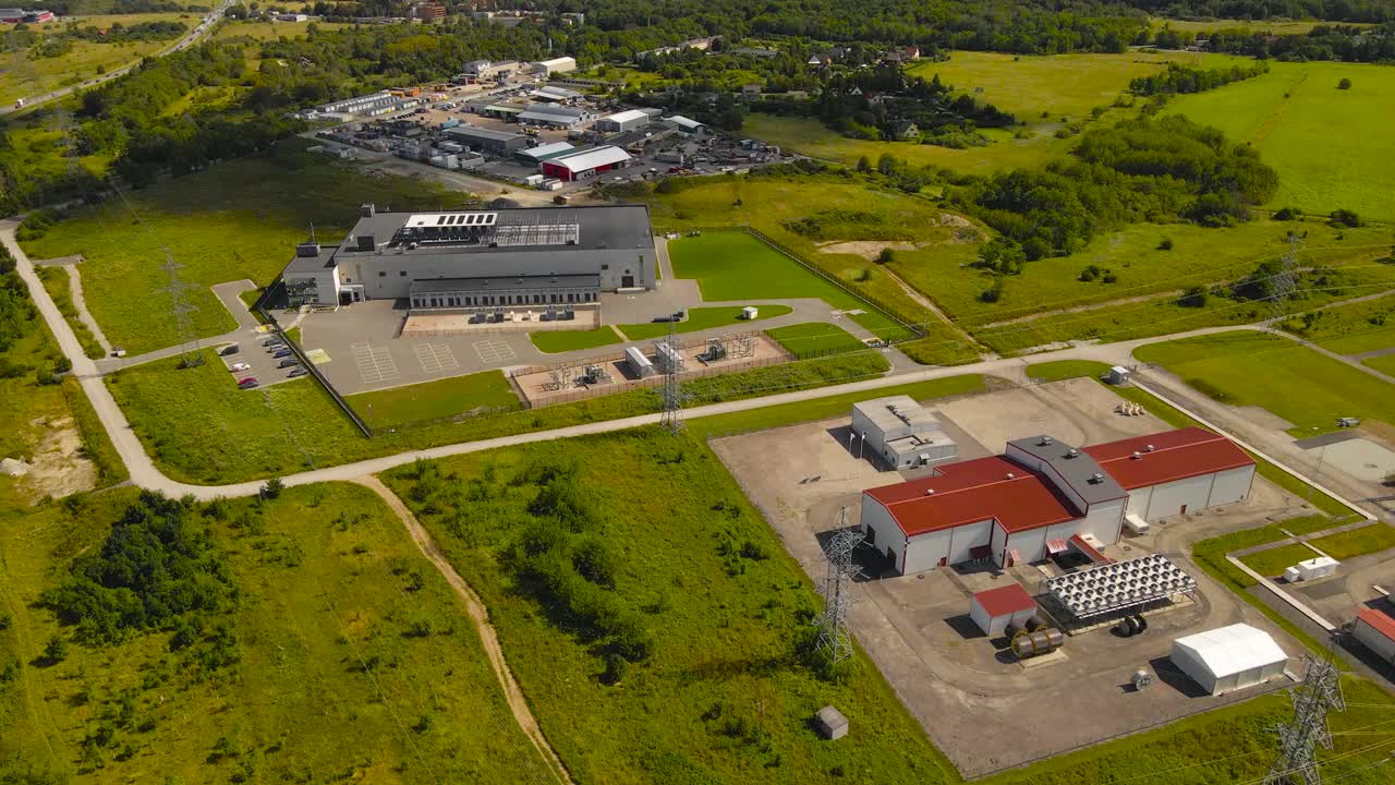 Aerial drone footage flying over a large server data center and an electrical power station powering the server housing at a grassy green countryside at summer time. Electrical lines visible below