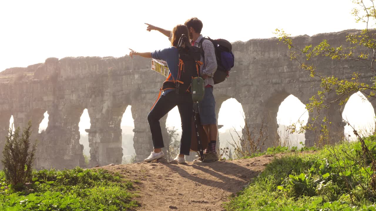 joven pareja encantadora mochileros turistas leyendo mapa señalando direcciones acueducto romano arcos en parco degli aquedotti parque ruinas en roma en romántico amanecer brumoso con guitarra y saco de dormir cámara lenta