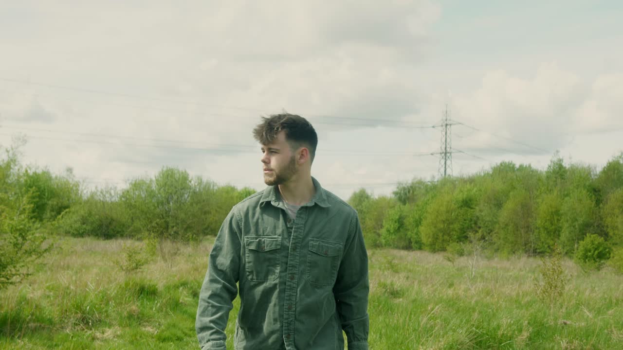 Close-up of young man standing still in grassy field looking calmly toward camera in moody daylight, walking into focus