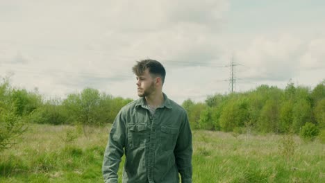 close-up of young man standing still in grassy field looking calmly toward camera in moody daylight, walking into focus