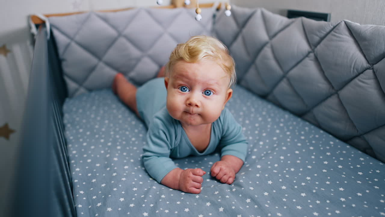 Caucasian infant with adorable plump cheeks lies peacefully in the crib. Lovely cute blue-eyed baby boy chewing something.