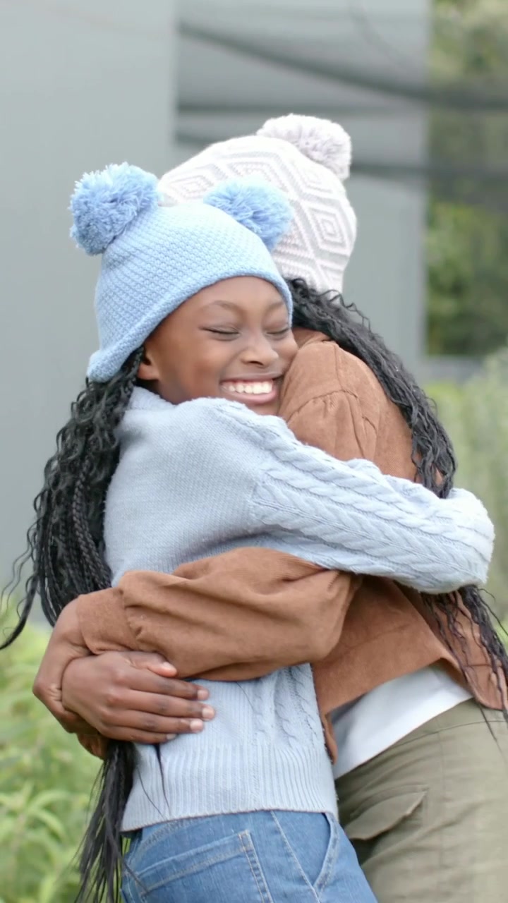 Vídeo vertical: Menina com gorro de tricô azul entrando e abraçando uma amiga no jardim, celebrando a amizade.