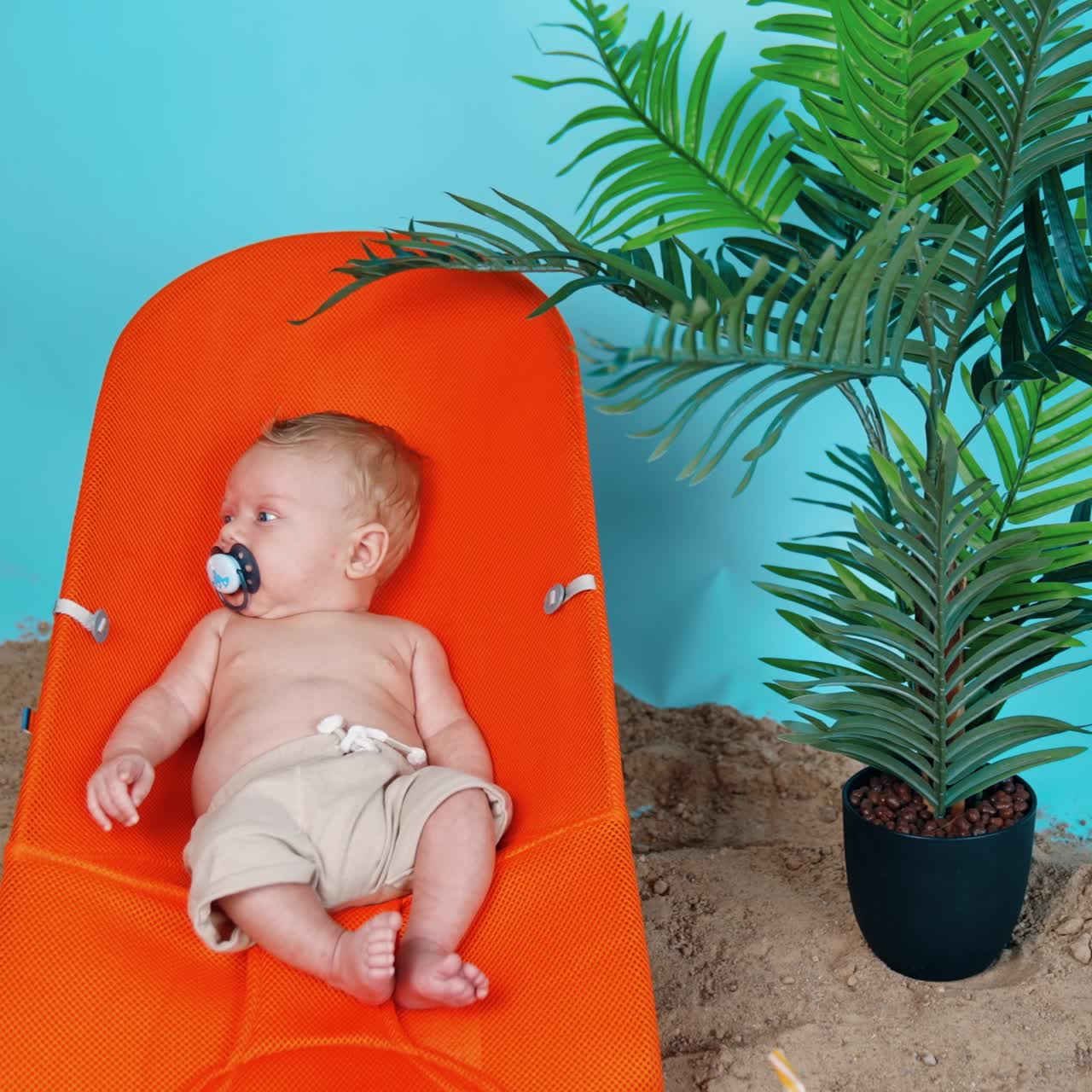 Sweet little baby in shorts and with pacifier lies on the orange chair. Beach sand and plant pot around the kid