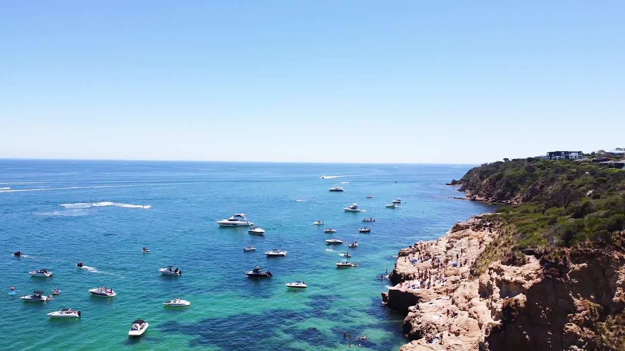toma aérea de botes y jetskis en el agua mientras la gente salta de las rocas