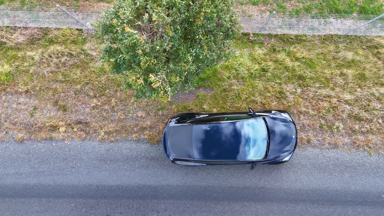 A dark vehicle travels along a quiet country road, passing a single tree, captured from directly overhead in bright natural daylight with smooth drone movement