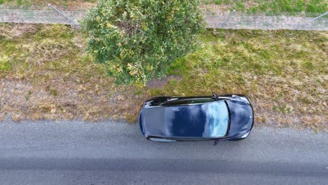 a dark vehicle travels along a quiet country road, passing a single tree, captured from directly overhead in bright natural daylight with smooth drone movement