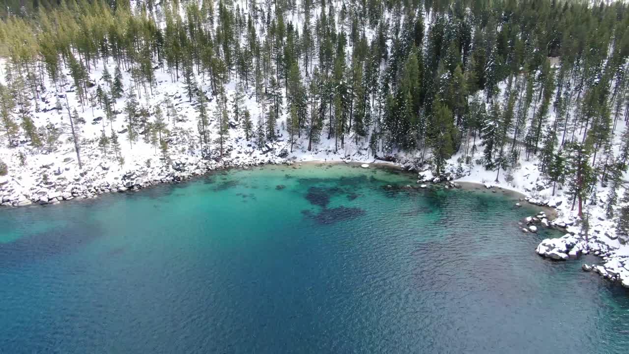 High-res 4K drone footage reveals clear waters of Lake Tahoe in a secret bay in California. A small cove lies on the east shore of the Sierra Nevadas in the mountains lined with trees and snow.