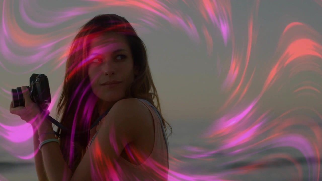 Donna in piedi sulla riva che tiene una macchina fotografica, guarda a sinistra, causando vortici rosa che le circondano la testa mentre cattura il tramonto