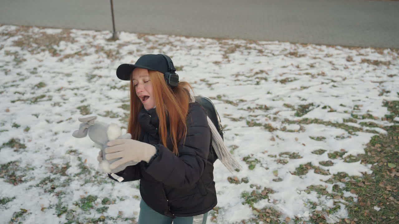 Woman holds pigeon in snowy park, redhead commuter with backpack and cap cradles small bird with gloved hands, headphones visible, suburban sidewalk dusted with snow, frames show passerby noticing