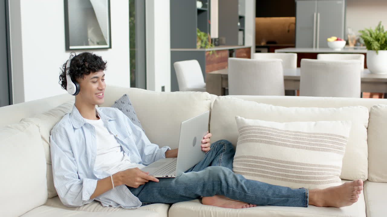 Relaxing on sofa, teenage boy using laptop and wearing headphones at home