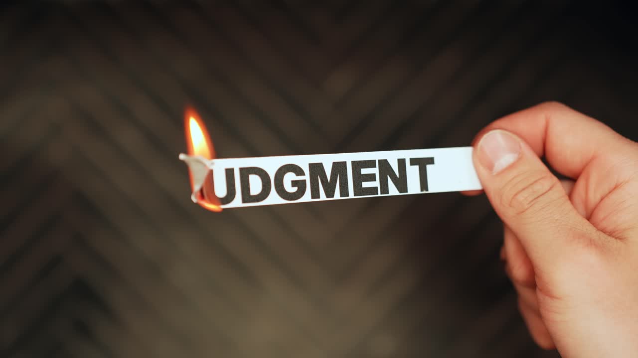 Close-up of a hand holding a paper strip labeled “JUDGMENT” as the edge burns with flame on a dark background. Letting go of criticism and shame concept with copy space