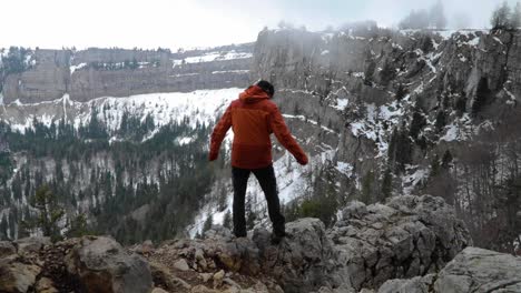 hombre parado en el borde del acantilado con vistas al paisaje invernal, levantando los brazos