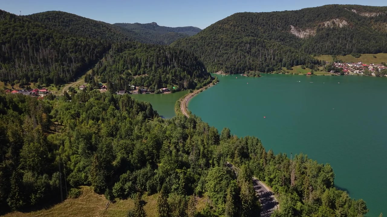 Een luchtbeeld toont een kronkelende weg die door een weelderig groen bos leidt naar een sereen bergmeer onder een helderblauwe hemel