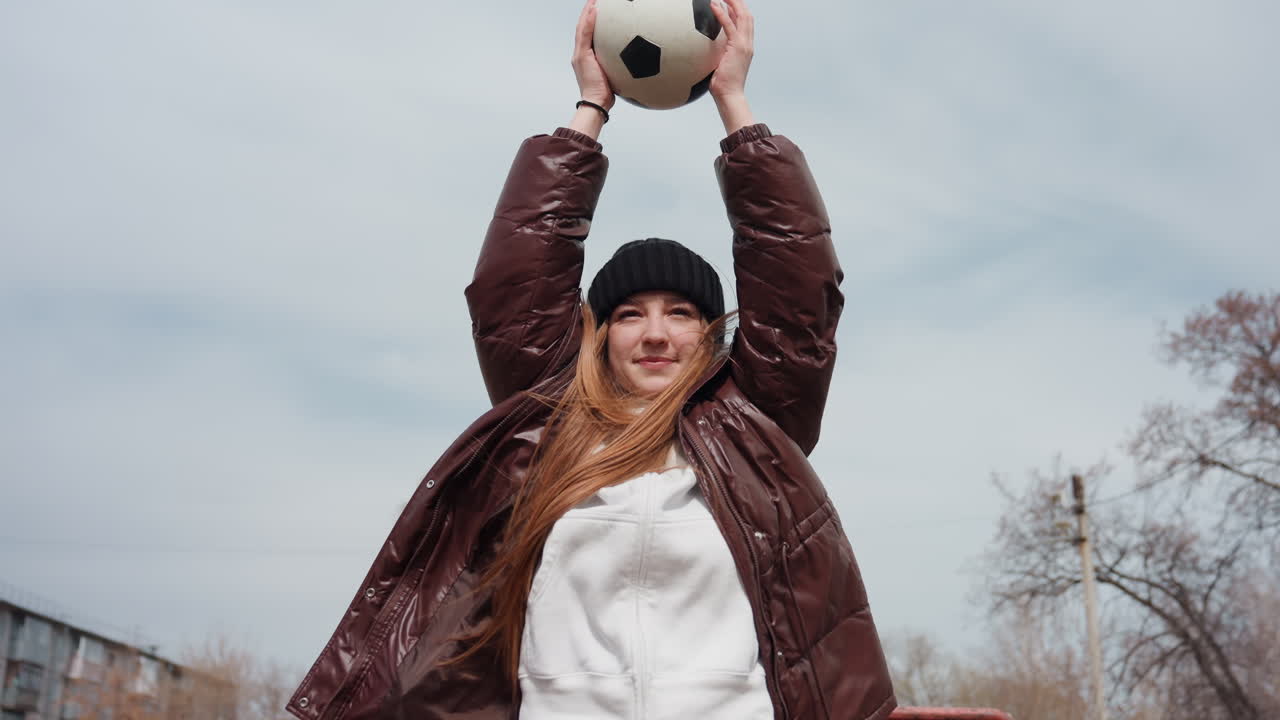 야외에서 재킷을 던지며 머리를 휘날리고 팔을 든 백인 십 대, 근심 걱정 없는 미소와 비니가 보임, 공원 같은 도시 필드에서 흐린 겨울 하늘을 배경으로 패딩 코트가 움직이는 모습, 활기차고 장난기 넘침