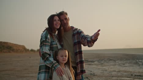 Un-Hombre-Moreno-Feliz-Con-Cabello-Gris-Con-Una-Camisa-A-Cuadros-Se-Para-Y-Disfruta-De-La-Vista-Con-Su-Esposa-Morena-Con-Una-Camisa-A-Cuadros-Verde-Y-Su-Pequeña-Hija-Rubia-Durante-Sus-Vacaciones-Fuera-De-La-Ciudad-En-Una-Playa-Desierta-En-La-Noche-En-Verano