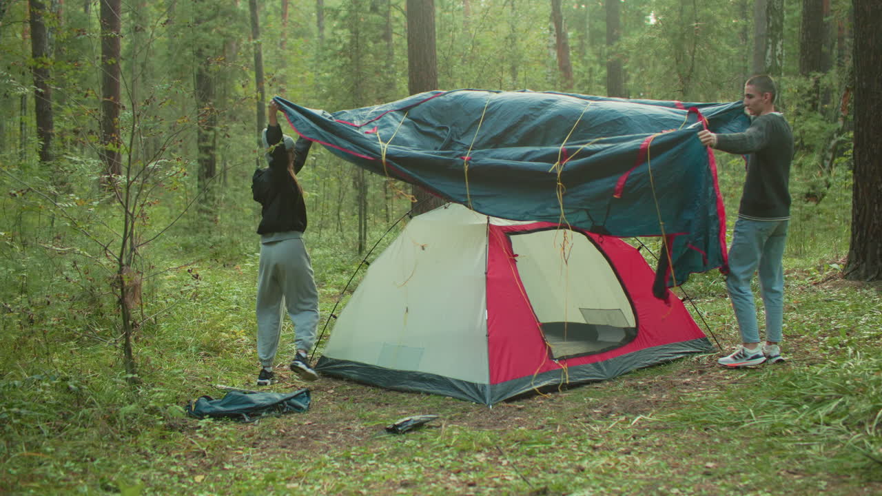 Couple lifts tent cover over pitched tent while camping in lush forest, coordinating setup effort in peaceful outdoor setting surrounded by trees, grass, and soft morning light