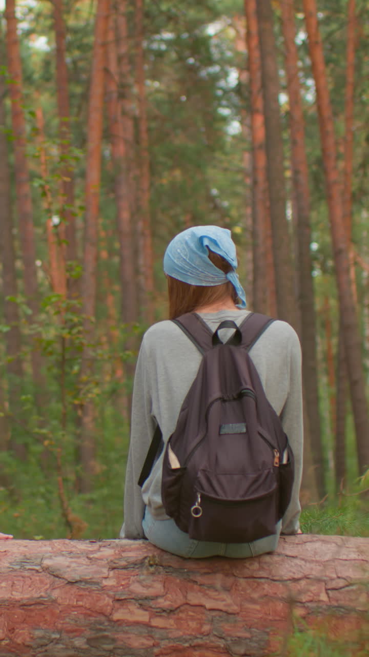 Hikers share warm moment on fallen tree in peaceful forest, one wearing blue bandana and black backpack smiles warmly, while her companion in green shirt with red bag cleans her hand after sitting