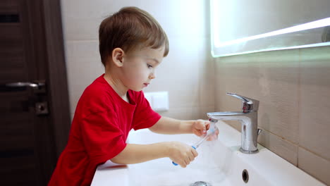 four-year-old toddler boy in red t-shirt washing a toothbrush. kid closes the tap and steps down from the sink.