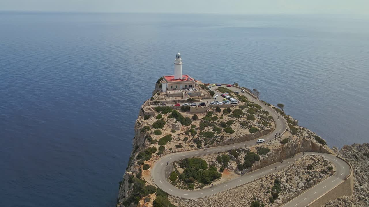 An aerial view of a lighthouse perched on a high rocky cliff, connected by a winding coastal road. The vast expanse of calm blue sea stretches endlessly into the horizon.