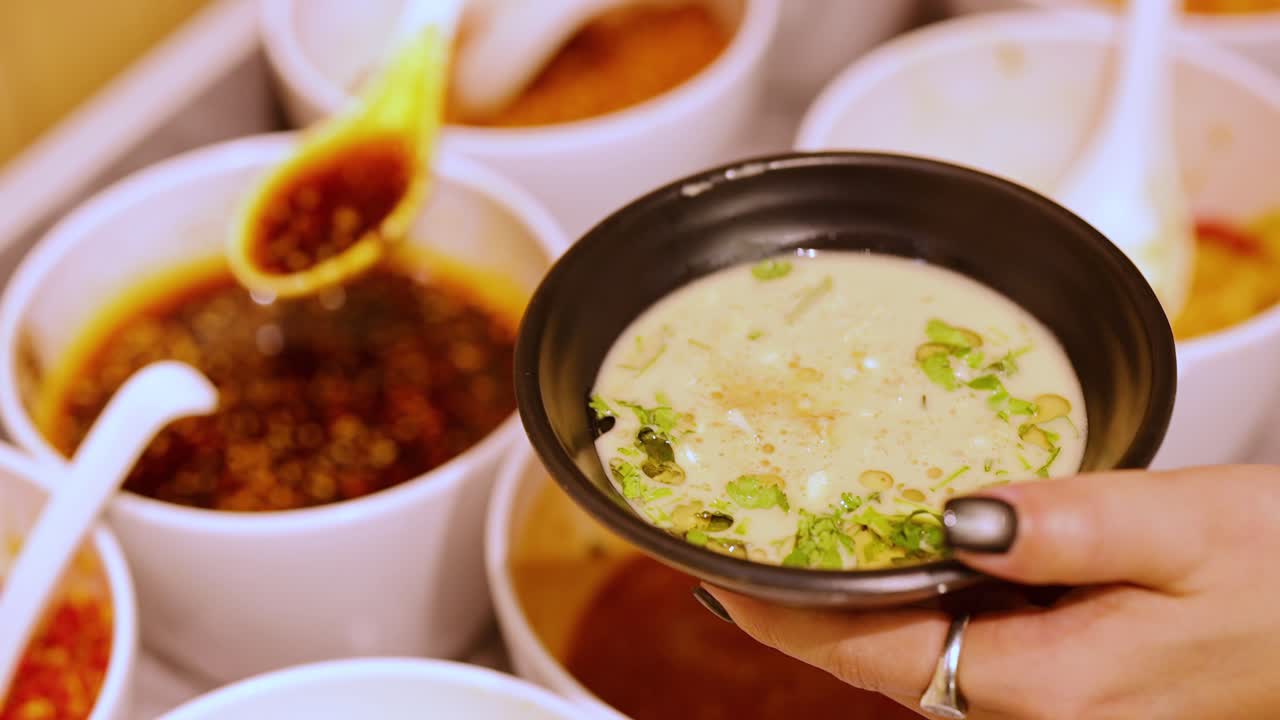 Hand pours chili oil onto sesame sauce at self-service condiment bar under warm indoor lighting