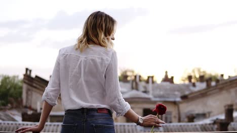 mulher sensual e magra de calças jeans e camisa branca está de pé em uma varanda com uma flor vermelha na mão. garota loira. bela perspectiva da cidade. traseira