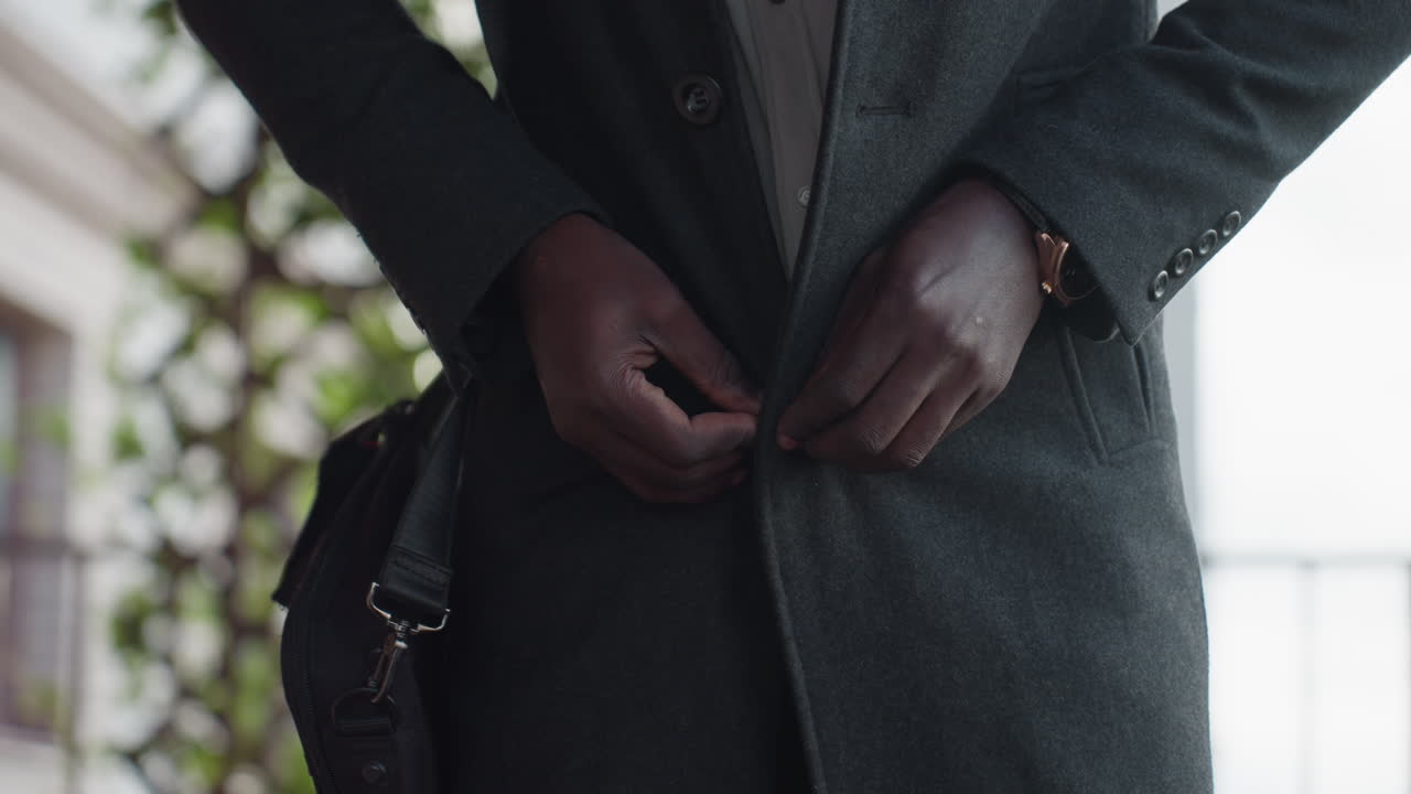 어두운 울 코트의 단추를 푸는 손, 바랜 흰색 셔츠와 손목시계, 노트북 가방 끈이 보이는 허리 높이의 클로즈업 뷰, 난간과 담쟁이가 있는 부드러운 배경, 텍스처와 미묘한 움직임에 초점이 맞춰져 있어요.