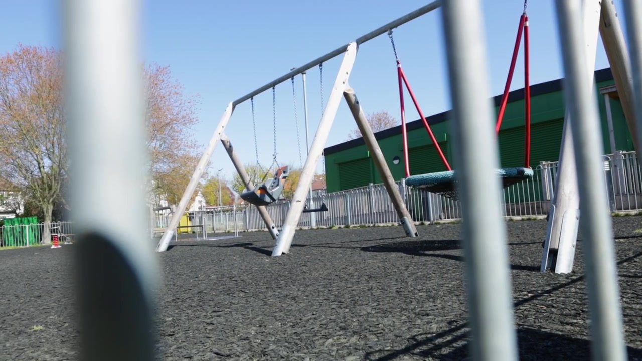 Columpios de parque infantil vacíos detrás de una valla metálica bajo un cielo azul despejado