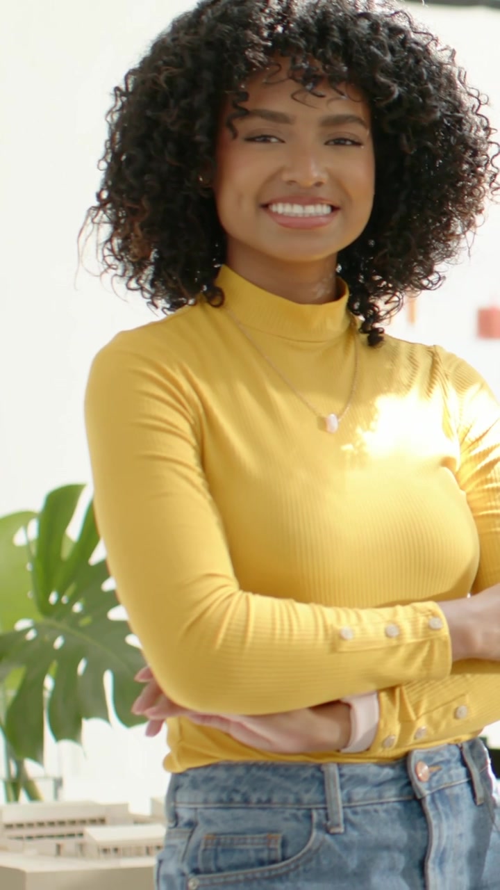 Vídeo vertical: Mulher sorrindo enquanto a câmera filma, levando a mão ao pescoço, cruzando os braços com blusa mostarda.