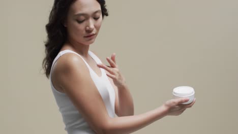 asian woman with dark hair applying cream on her skin on beige background, copy space, slow motion