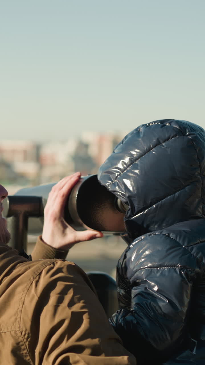 un padre sostiene a su hijo hasta un telescopio, guiándolo mira a través de él, el padre sonríe, asegurando una vista estable mientras el paisaje de la ciudad se extiende en el fondo