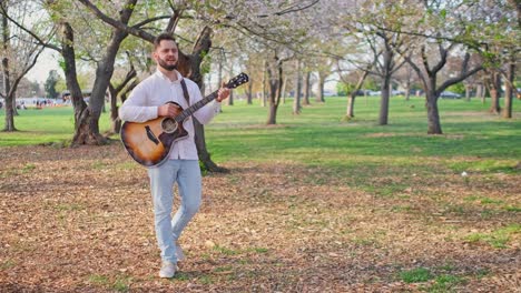 Adult-man-and-musician,-singer,-guitarist,-and-artist-is-walking-through-a-park-singing-a-song-and-strumming-his-guitar