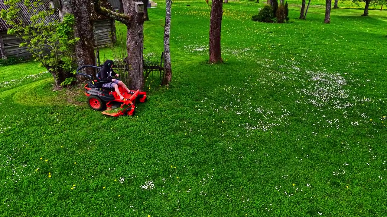 Flygfoto som sänker sig över gröna träd och avslöjar en man som klipper gräs på en röd åkgräsklippare vid en trästuga i en lantlig by i Lettland.