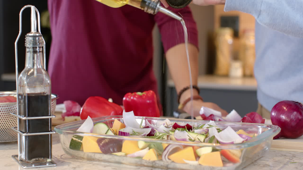 Pouring olive oil over vegetables, multiracial gay couple preparing meal in kitchen, at home