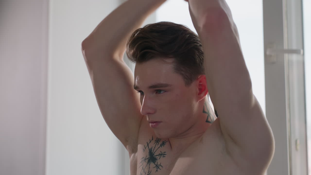 focused gym lover stretches resistance band overhead near window with sunlight highlighting toned physique, displaying chest tattoo and determined expression in calm indoor setting