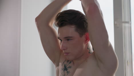 focused gym lover stretches resistance band overhead near window with sunlight highlighting toned physique, displaying chest tattoo and determined expression in calm indoor setting