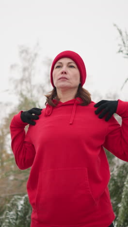 lady performing hand stretch by turning arm in fitness routine outdoors, dressed in athletic wear, background featuring frosted tree, bag on bench, snow covering ground