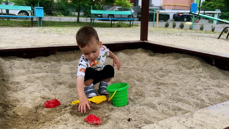 beautiful child sitting in the sandbox grabs some sand and puts it into bucket. cute toddler playing in the sandpit at playground.
