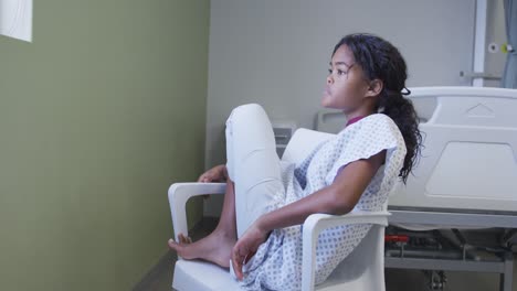 mixed race girl sitting on chair in hospital patient room looking through the window