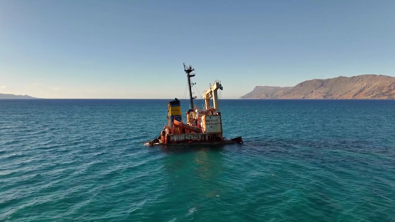 Il relitto della Manassa Rose M giace al largo nella baia di Kissamos, vicino a Drapanias, Kissamos, Chania, a Creta, con la sua sovrastruttura arrugginita che emerge dalle acque limpide dell'Egeo e montagne aspre sullo sfondo, ripresa aerea con drone in allontanamento