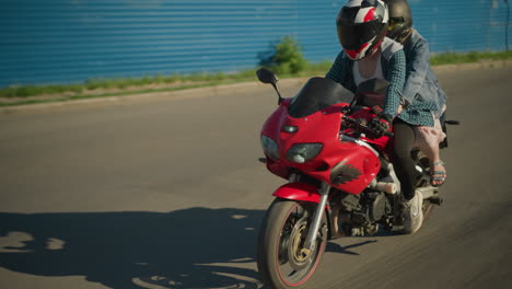 two ladies are riding a red power bike, both wearing helmets, as they travel through an urban area, their shadows are cast on the ground as they maintain focus while moving forward