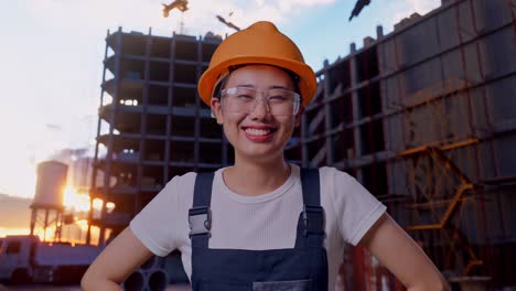 close up of asian woman worker wearing goggles and safety helmet standing with arms akimbo looking at camera and smiling at construction site
