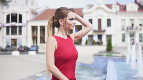 Mujer-Hermosa-Y-Relajada-Con-Cabello-Largo-Y-Rubio-Sentada-Junto-A-La-Fuente-De-La-Ciudad-Y-Corrigiendo-Su-Peinado.-Mujer-En-Vestido-Rojo